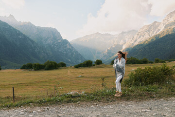 Fototapeta premium A solitary woman stands in an open field with dramatic mountains in the distance, wearing casual outerwear and a relaxed smile, exuding calm adventure.