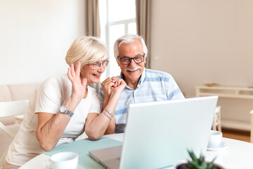 Fototapeta premium Senior couple video call. Elderly people using laptop computer, communication and technology concept. Happy mature grandparents waving hand at screen, sitting at the table in living room.