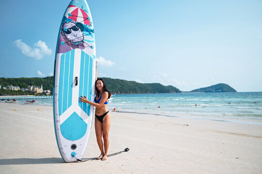 Happy Asian woman posing with a stand-up paddleboard on a sunny tropical beach