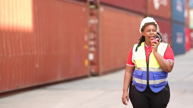 Black female worker talks on walkie talkie while walking through container yard with serious focus, showing logistics communication, safety checks, cargo inspection, port workflow.