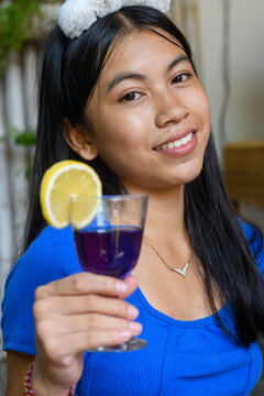 Close-up portrait of smiling Asian woman holding glass of butterfly pea blue tea with lemon slice