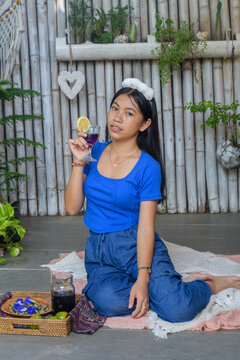Young Asian woman in blue casual wear sitting cross-legged with butterfly pea tea looking away thoughtfully