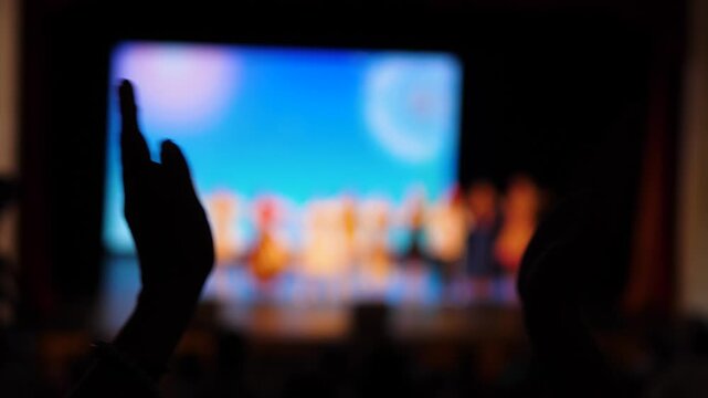 Silhouette of an audience member's hands enthusiastically clapping in the darkness of a concert hall. In the blurred background, actors and dancers bow on a brightly lit stage after the performance.