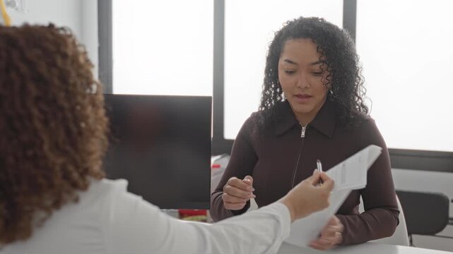 Woman signing medical consent form with right hand and pen at clinic desk in building while reviewing paperwork and interacting with a clinician; trust professionalism.
