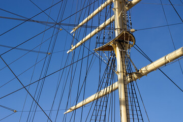 Fototapeta premium Tall ship mast with rigging and sails against a clear blue sky, showcasing intricate details of ropes and wooden structure in a nautical setting