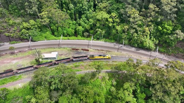 Aerial drone view of freight train moving slowly along railway tracks through dense forest landscape showing rail transport and cargo movement.