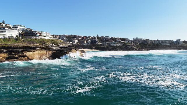 Ocean waves rolling over rocky shoreline with coastal houses overlooking water under clear sky forming a scenic seaside landscape in Bronte Beach Australia