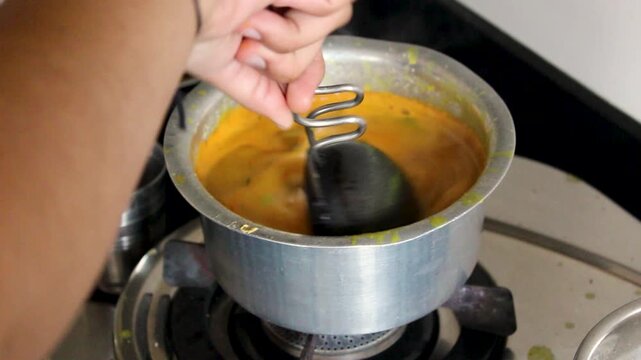 Hand stirring vibrant golden dal after adding tadka in a steel pot on gas stove. Ideal for Indian recipe videos, food documentaries, and lentil curry cooking content.