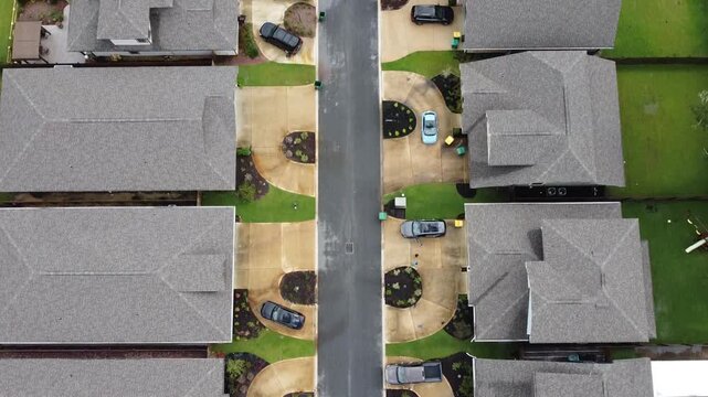 Aerial top view of suburban residential street with houses aligned on both sides showing driveways parked vehicles and neat landscaping in a structured neighborhood layout