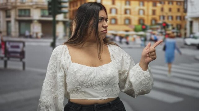 Woman in white embroidered blouse with hands raised and pointing finger while frowning at traffic in a busy city street; frustration.