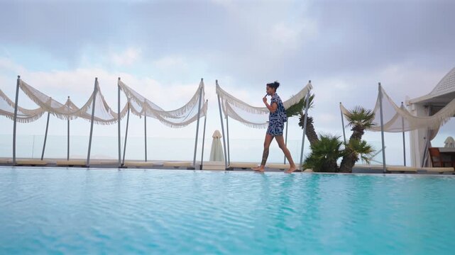 Young Indian woman in blue dress walks barefoot along the edge of turquoise infinity pool at luxury resort in Santorini Greece. Decorative canopies and palm trees frame the overcast sky