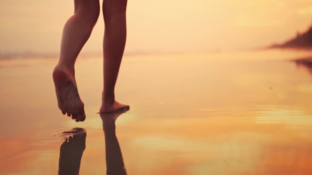 Close up of bare feet of young Asian woman walking on wet sand beach at sunset, lower body part of female strolling on sea shore during golden hour, tropical summer vacation, travel lifestyle.