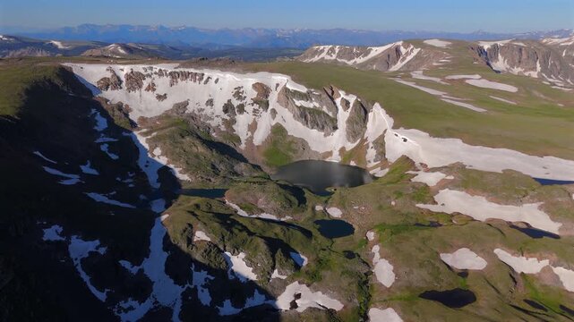 Aerial drone footage soars over stunning alpine landscape featuring glacial lakes nestled in rugged mountain terrain with patches of melting snow, green tundra meadows.
