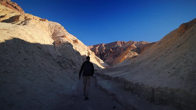 Male hiker with backpack walks through Golden canyon, Death Valley with cream-colored eroded walls and distant red rock formations bathed in warm golden hour light under blue cloudless sky in Death.