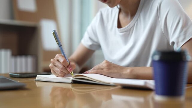 Focused student or female entrepreneur writing in a notebook while sitting at a desk with a coffee cup, planning her schedule, taking notes, or studying for an exam