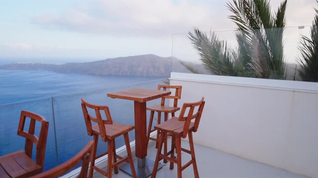 Elegant wooden bar furniture on white terrace with glass railing offers stunning view of calm Aegean Sea and volcanic caldera cliffs in Santorini Greece under overcast sky with palm fronds swaying.