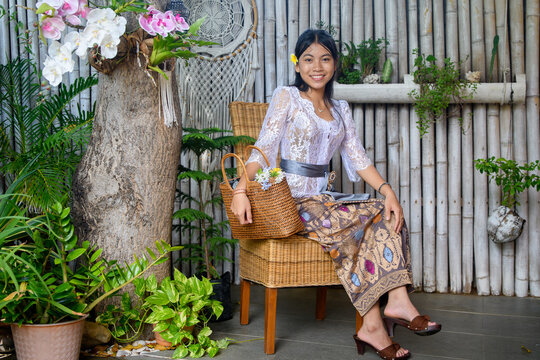 Happy Balinese woman in lace kebaya and ikat batik skirt posing on rattan furniture in tropical boho garden with orchids