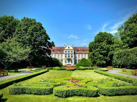 Abbot's Palace in Oliwa Park, Gdansk, Poland, features a historical building with a Baroque architectural style. The palace has a symmetrical facade with a red-tiled roof and pastel-colored walls. 