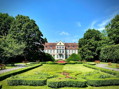 Abbot's Palace in Oliwa Park, Gdansk, Poland, features a historical building with a Baroque architectural style. The palace has a symmetrical facade with a red-tiled roof and pastel-colored walls. 