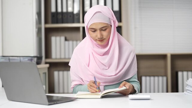 Young muslim businesswoman wearing a pink hijab writing in her agenda and organizing her schedule at her desk while working in a modern corporate office environment