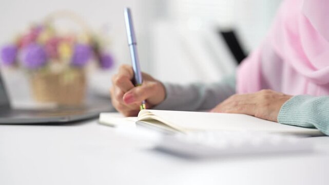 Muslim woman wearing a hijab writing in a notebook with a fountain pen at her desk, focusing on her hands and the creative process of journaling or studying in a modern workplace