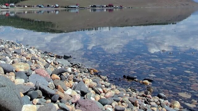 Experience the serene clarity of Khuvsgul Lake in Mongolia. Brilliant pebbles line the shore while sky and distant landscape mirrors perfectly on the calm, pristine water surface, a tranquil moment.