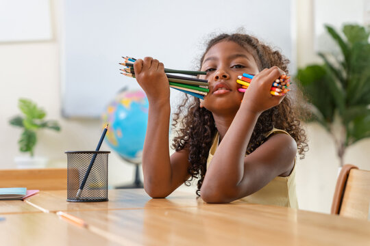 Portrait of a happy little girl learning in an elementary school classroom, Cute African American girl holding colored pencils in a classroom, Young student playing with colorful pencils at desk