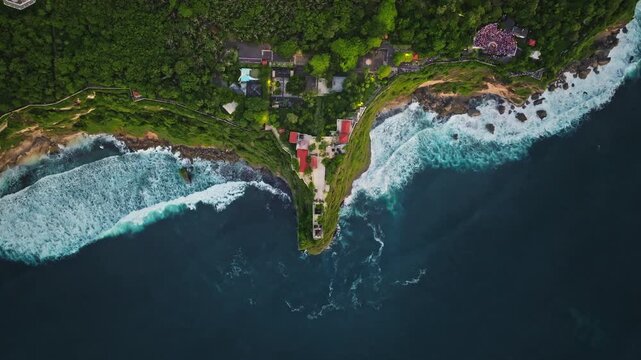 Bali, Uluwatu Temple perched on a high cliff overlooking the Indian Ocean, lush green vegetation lining coast and powerful ocean waves crashing against rocky shoreline below. Aerial view drone flight