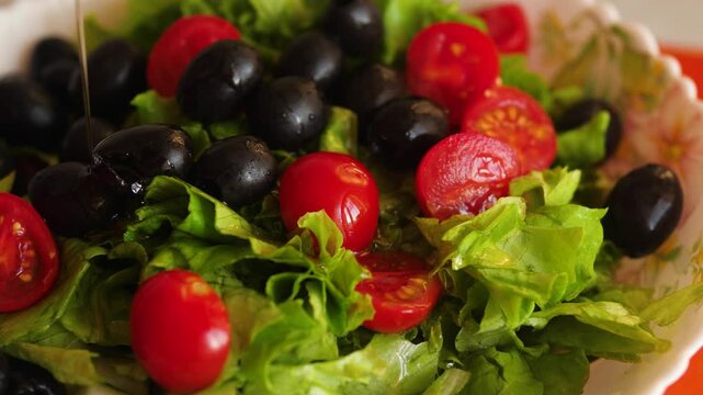 Fresh Greek salad with cherry tomatoes, black olives and lettuce leaves being drizzled with dressing. A thin stream of olive oil or sauce pours onto the appetizing juicy vegetables in a bowl.