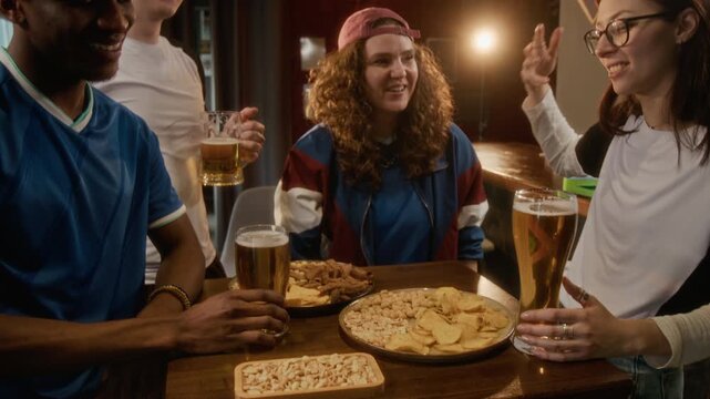 Handheld shot of joyful young multiethnic friends in team apparel raising arms and bumping fists while enjoying beer and snacks in sports bar