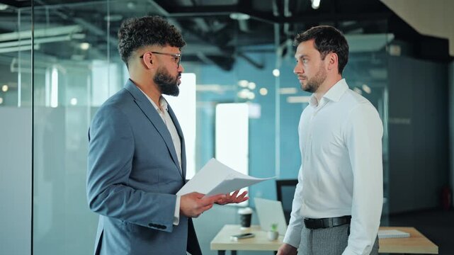 Annoyed corporate boss with paper document expressing dissatisfaction during project review. Employee in white shirt feeling nervous about bad report in glass office.