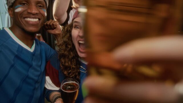UGC shot of excited young diverse friends wearing team jersey cheering with foam finger while looking at camera, celebrating joyous victory in sports bar together