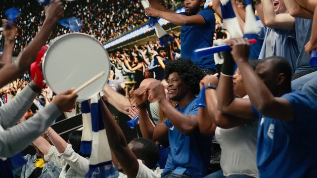 Dynamic football crowd leader motivating blue supporters with megaphone and drum during coordinated chant in packed stadium stands.