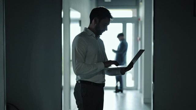 Serious business professional working on laptop in dim office hallway during blackout. Corporate employee standing in dark corridor while coworker stands in background during electricity outage.