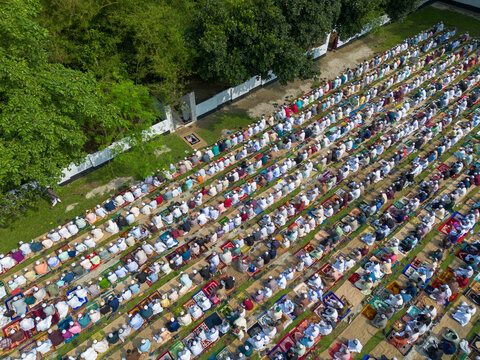 Joypurhat, Bangladesh - 28 May 2026: Aerial view of a large congregation of Muslim men performing Eid prayer in neat rows on colorful mats outdoors surrounded by lush green trees.