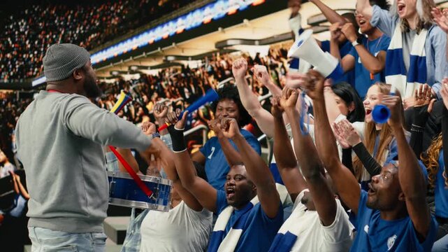 Dedicated football fan leader directing blue supporters with drum and megaphone in stadium terrace during loud coordinated soccer chant.
