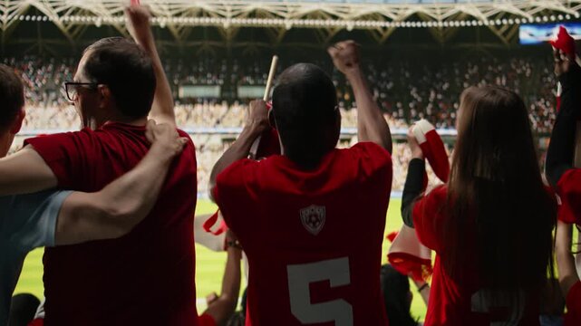 Back view of passionate soccer supporters in red jerseys cheering in packed stadium during live match, authentic crowd with raised scarves, chanting and loud stadium atmosphere for team spirit.