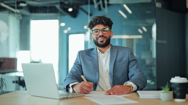 Confident Middle Eastern businessman working with laptop and writing on paper at office desk. Arab executive posing for camera at contemporary corporate workplace.