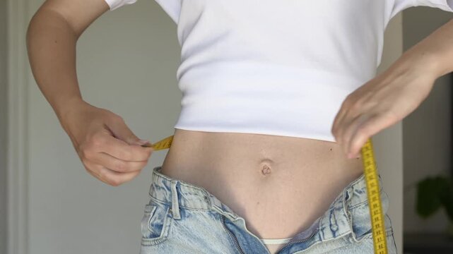 Woman measures waist with tape measure next to open jeans showing diet and weight tracking progress closeup. Measurement footage suits weight loss clinic promotion, slimming medication, diet program