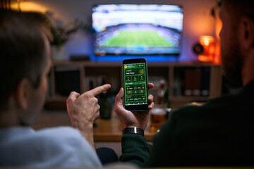 Obraz na płótnie Canvas Young adult men watching football match and checking betting app on smartphone, expressing male friendship and shared leisure. Useful for sports betting, gaming, entertainment marketing