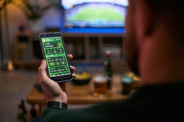 Young adult man holding smartphone, checking sports betting app while watching match with friend,...