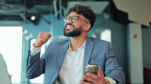 Joyful Middle Eastern businessman in glasses looking at phone screen, laughing genuinely and celebrating business win. Enthusiastic corporate worker holding smartphone at modern workspace desk.
