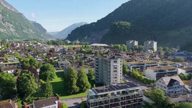 Aerial drone view of Glarus, Switzerland, at sunrise, showing residential buildings, apartment blocks, green spaces, and surrounding alpine mountains under a clear sky.