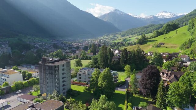 Aerial drone view of Glarus, Switzerland, at sunrise, showing residential houses, green fields, and alpine mountains in soft morning light.