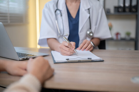 Close up of woman doctor writing in clipboard talking to patient at wooden table in hospital clinic.
