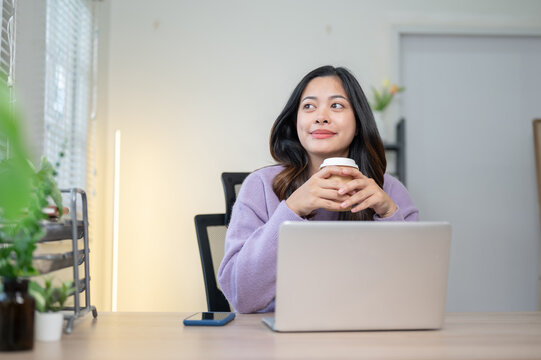 Pretty asian woman holding coffee looking away from laptop sitting at wooden table in home office