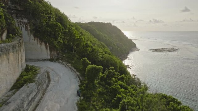 Unpaved road leading to lush green cliffside towards the calm ocean and Green Bowl Beach, showcasing the natural coastal landscape and limestone formations of Bali, Indonesia. Aerial view drone flight