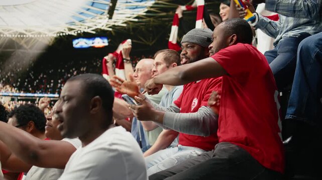 Diverse soccer fans following in-play live odds on phone betting app, cheering and watching exciting football match from stadium stands.