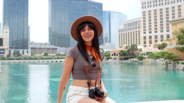 Cheerful female traveler wearing a hat sitting on a ledge with the iconic las vegas cityscape and a lake in the background, enjoying her summer vacation and smiling at the camera