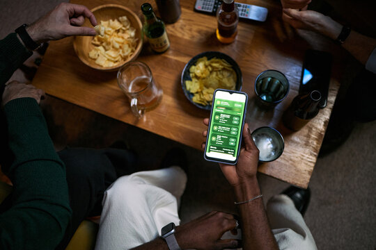 Overhead shot of young male friends gathering around table, checking smartphone app, sharing drinks and snacks, representing social connection and digital lifestyle. Friendship, leisure, app marketing